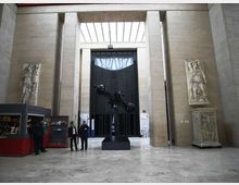 Ampio atrio museale con pareti in pietra chiara e soffitto alto con lucernario, dominato da un grande portale scuro sul fondo. Al centro si trova una scultura metallica su basamento, affiancata da grandi rilievi e statue in pietra lungo le pareti.