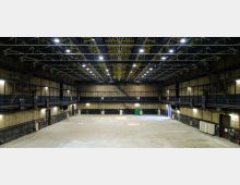 Grande capannone industriale vuoto con ampia pavimentazione in cemento e pareti scure, illuminato da numerosi faretti a soffitto. Struttura metallica a traliccio con passerelle laterali sopraelevate e porte di servizio lungo i lati.