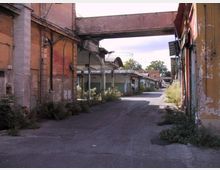 Strada asfaltata in un complesso industriale dismesso, fiancheggiata da edifici bassi con pareti scrostate e capannoni con tettoie in metallo. Un passaggio sopraelevato collega due strutture sopra la carreggiata, con vegetazione incolta lungo i bordi.