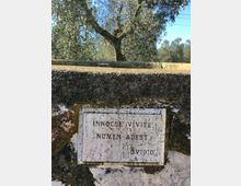 Muro in pietra intonacata e invecchiata con una targa rettangolare in marmo incassata, incisa con una scritta in latino. Sullo sfondo si vedono alberi, tra cui un ulivo, sotto un cielo azzurro.