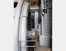 Interno di un impianto industriale o museo tecnico con grandi macchinari metallici grigi affiancati, tubazioni e componenti meccanici visibili. In primo piano si nota una ringhiera di sicurezza lungo un passaggio stretto tra le strutture.