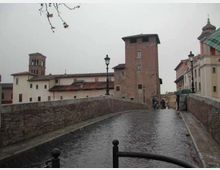 Ponte pedonale in pietra con pavimentazione in ciottoli e parapetti in muratura, che conduce verso edifici storici con torri in mattoni. Lungo il ponte si vedono lampioni e, sullo sfondo, un complesso di case e strutture d’epoca sotto un cielo grigio.