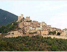 Borgo medievale in pietra arroccato su una collina, con case addossate tra loro e una torre o castello sulla sommità. Sullo sfondo si vedono un pendio montuoso e, in primo piano, una fascia di vegetazione e alberi.