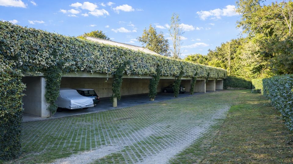 Parcheggio coperto all’aperto con una lunga tettoia in cemento sostenuta da pilastri, ricoperta di edera e con posti auto in ombra. Davanti c’è un vialetto pavimentato con griglie erbose, circondato da siepi e alberi sotto un cielo azzurro.