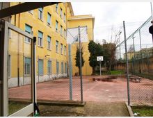 Cortile esterno recintato con campo da basket in cemento rosso, canestro e panchina, affiancato a un grande edificio giallo a più piani con finestre e grate. Sullo sfondo si vedono alberi e una piccola area verde oltre la rete metallica.