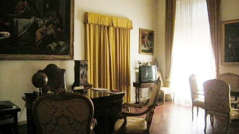 Salotto in stile classico con pavimento in legno, mobili d’epoca e poltrone imbottite disposte attorno a un tavolino. Alle pareti sono presenti grandi quadri e tende pesanti incorniciano una finestra alta e luminosa con portafinestra.