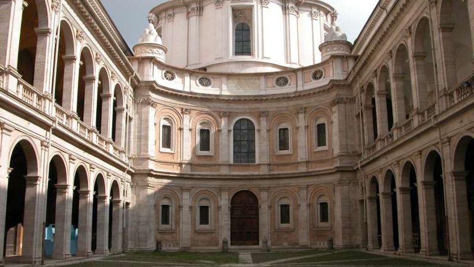 Cortile interno di un complesso storico con porticati ad archi su due livelli lungo i lati e, sul fondo, una facciata curvilinea con finestre e grande portale, sormontata da una cupola. Pavimentazione con disegni geometrici in pietra e aree verdi.