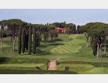 Campo da golf con ampi fairway ondulati a strisce, delimitato da cipressi e pini mediterranei. In primo piano si vede un piccolo ponte sul percorso e sullo sfondo un edificio con tetto rosso tra gli alberi.