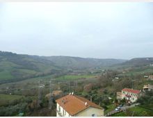 Vista panoramica di una valle collinare con campi verdi e boschetti, sotto un cielo nuvoloso. In primo piano si vedono case con tetti in tegole rosse, una strada e tralicci dell’alta tensione che attraversano il paesaggio.