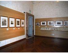 Interno di una sala espositiva con pareti chiare e pannelli in legno che ospitano fotografie incorniciate e didascalie. Pavimento in parquet scuro e porta centrale con cornice in pietra, con una lunga vetrina orizzontale a parete che espone materiali e immagini.