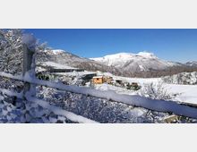 Paesaggio montano innevato con catena di montagne sullo sfondo sotto un cielo limpido. In primo piano una recinzione e alberi coperti di neve, e nella valle alcuni edifici con tetti innevati.