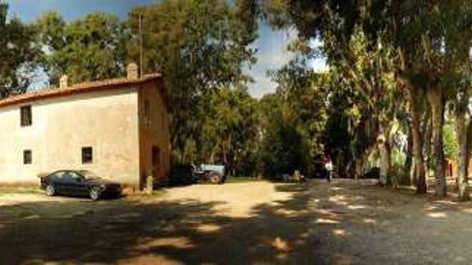 Cortile sterrato ombreggiato da alti alberi, con una piccola casa rurale intonacata al centro e altri edifici bassi sullo sfondo. Sul lato sinistro si vede un muro rosso con un arco d’ingresso e, nell’area, alcuni veicoli parcheggiati lungo i bordi del piazzale.