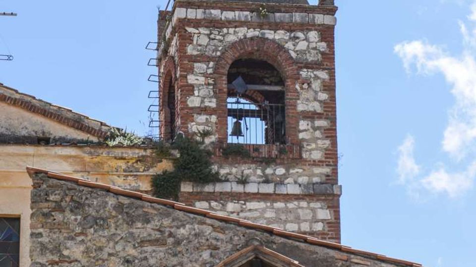 Campanile di una chiesa in muratura mista di pietra e mattoni, con aperture ad arco e campane visibili, sormontato da una guglia con croce. In primo piano si vede il tetto in tegole e una nicchia con tondo decorativo sulla facciata, sotto un cielo azzurro.