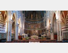 Interno di una chiesa con navata centrale e arcate a tutto sesto su colonne, pareti a fasce orizzontali e soffitto a volta. In fondo si vede il presbiterio rialzato con altare e grande abside decorata da affreschi e mosaici.