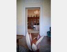 Vista attraverso una porta interna di un salone classico con pareti chiare modanate e pavimento in parquet, che conduce a una sala da pranzo. Nella stanza sullo sfondo si vedono un lampadario in cristallo, una grande credenza in legno con stoviglie esposte e un tavolo rotondo coperto da una tovaglia.