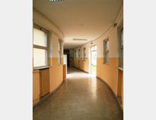 Corridoio interno curvo di un edificio pubblico, con pareti bicolori crema e arancione, pavimento in piastrelle grigie e finestre rettangolari lungo entrambi i lati. Illuminazione a plafoniere sul soffitto e una porta aperta in fondo che lascia entrare luce dall’esterno.