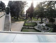 Giardino storico con vialetti in ghiaia e aiuole geometriche delimitate da siepi, circondato da alberi alti. A sinistra una lunga scalinata con balaustra e lampioni, a destra una fontana circolare con una scultura.