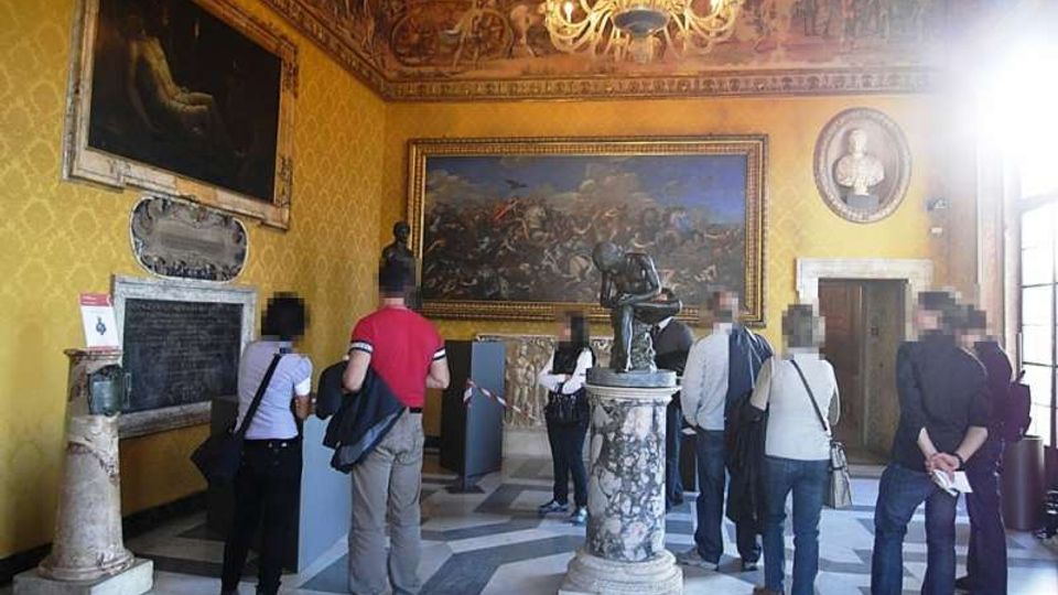 Sala museale con pareti giallo ocra decorate, grandi dipinti incorniciati e fregi affrescati vicino al soffitto, illuminata da un lampadario centrale. Al centro della stanza c’è una scultura su piedistallo in marmo e il pavimento è a motivi geometrici in pietra.