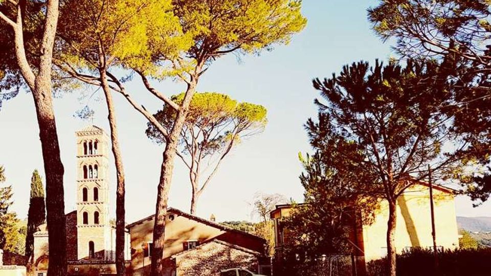 Veduta di un complesso storico con campanile in pietra a più ordini di bifore, affiancato da edifici bassi con tetti in coppi. In primo piano un’area alberata con alti pini mediterranei che incorniciano la scena.