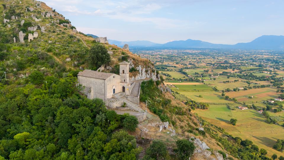 Piccola chiesa in pietra con campanile, costruita su un crinale roccioso coperto di vegetazione e raggiunta da un sentiero con muretti e scalinate. Sullo sfondo si apre una vasta pianura agricola con campi e paesi, con montagne all’orizzonte sotto un cielo sereno.