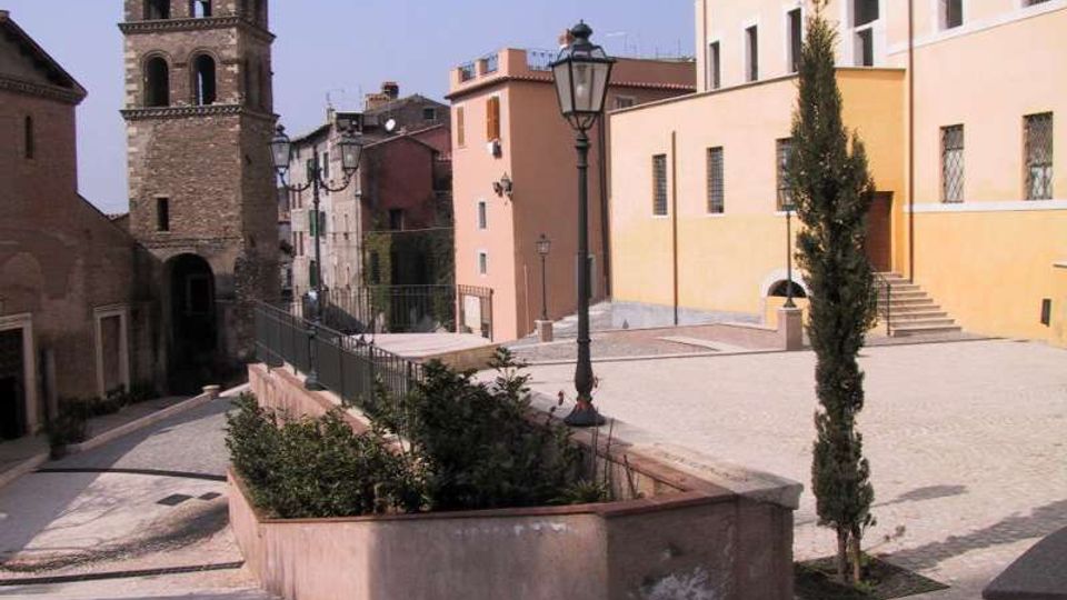 Piazza in pendenza di un borgo storico con pavimentazione in pietra, aiuole e lampioni in ferro battuto. Sullo sfondo si vedono una torre campanaria in pietra e facciate di edifici intonacati con finestre e scale esterne.