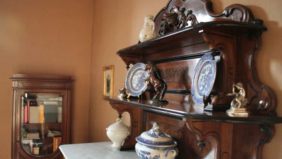 Sala d’interni con mobili in legno scuro in stile classico, tra cui una credenza con ripiani decorati con piatti in ceramica e statuette e un mobile vetrina nell’angolo. In primo piano si vede un tavolo con piano in marmo e un vaso in ceramica blu e bianca.