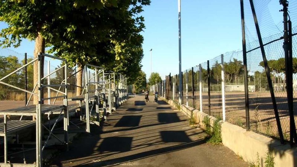 Viale asfaltato all’interno di un impianto sportivo, con alberi a sinistra e gradinate metalliche lungo il percorso. A destra si vedono recinzioni alte in rete e un campo sportivo in terra battuta, illuminato da un palo luce.