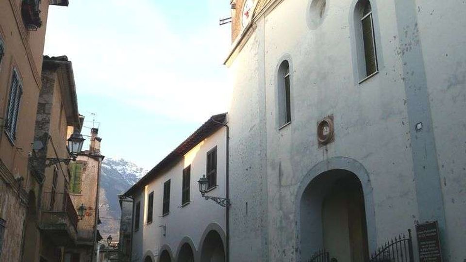 Strada stretta di un centro storico con una chiesa dalla facciata intonacata bianca, portale ad arco e finestre alte ad arco, affiancata da un portico con arcate. Sullo sfondo si vede un campanile in muratura e edifici storici con balconi e lampioni, sotto un cielo sereno.