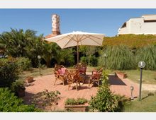 Patio esterno pavimentato in mattoni con tavolo e sedie in legno sotto un grande ombrellone bianco, circondato da vasi e lampioni da giardino. Sullo sfondo si vedono prato e vegetazione fitta con palme e un edificio chiaro con camino.