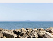 Costa marina con una barriera di grandi massi e blocchi di cemento in primo piano, affacciata su un mare calmo con orizzonte netto. In lontananza si intravede una piccola isola o promontorio sullo sfondo sotto un cielo sereno.
