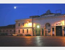 Facciata di una stazione ferroviaria storica illuminata di sera, con grandi archi e colonne e l’insegna “METRO” su un ingresso laterale. Davanti si apre una piazza lastricata con alcune auto parcheggiate sotto un cielo blu scuro.