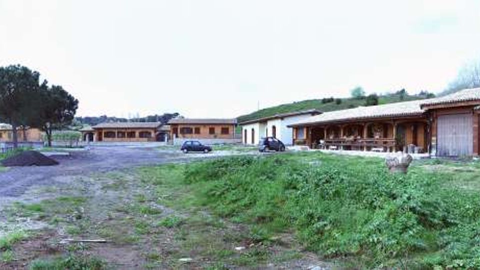 Ampio piazzale sterrato in una zona rurale, circondato da prati e leggere colline, con diversi edifici bassi e lunghi in legno e muratura. In primo piano si vede una strada in ghiaia e, sullo sfondo, tettoie aperte con cataste di legna e qualche veicolo parcheggiato.