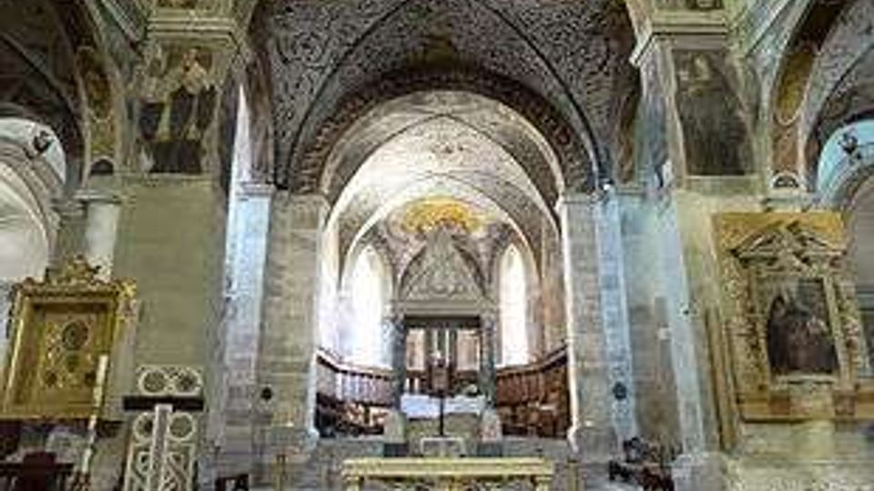 Interno di una chiesa storica con navate in pietra, archi a tutto sesto e volte affrescate; al centro si vede l’altare e, sullo sfondo, l’abside illuminata. Ai lati sono presenti cappelle e monumenti lapidei, con pavimento in lastre e un percorso centrale verso il presbiterio.