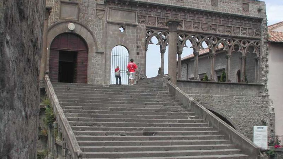 Ampia scalinata in pietra che conduce all’ingresso ad arco di un edificio storico fortificato, con muri in blocchi di pietra. Sul lato destro si estende una loggia con archi gotici traforati affacciata su un cortile interno.