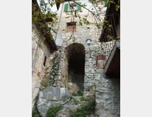 Cortile stretto in un borgo con edifici in pietra, un grande arco che conduce a una scalinata e muri con finestre e persiane verdi. In primo piano si vedono roccia e vegetazione, con rami di albero sopra e un lampione fissato alla parete.