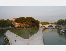 Veduta di un’isola fluviale con una passeggiata in pietra che segue la riva, alberi fitti e edifici storici. Sullo sfondo si vede un ponte in muratura a più arcate che attraversa il fiume.