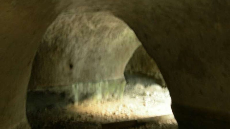 Interno di una piccola galleria sotterranea in pietra con volta ad arco, pareti grezze e pavimento in terra e ghiaia. Una zona illuminata in fondo mette in evidenza depositi e macchie di umidità lungo le pareti.