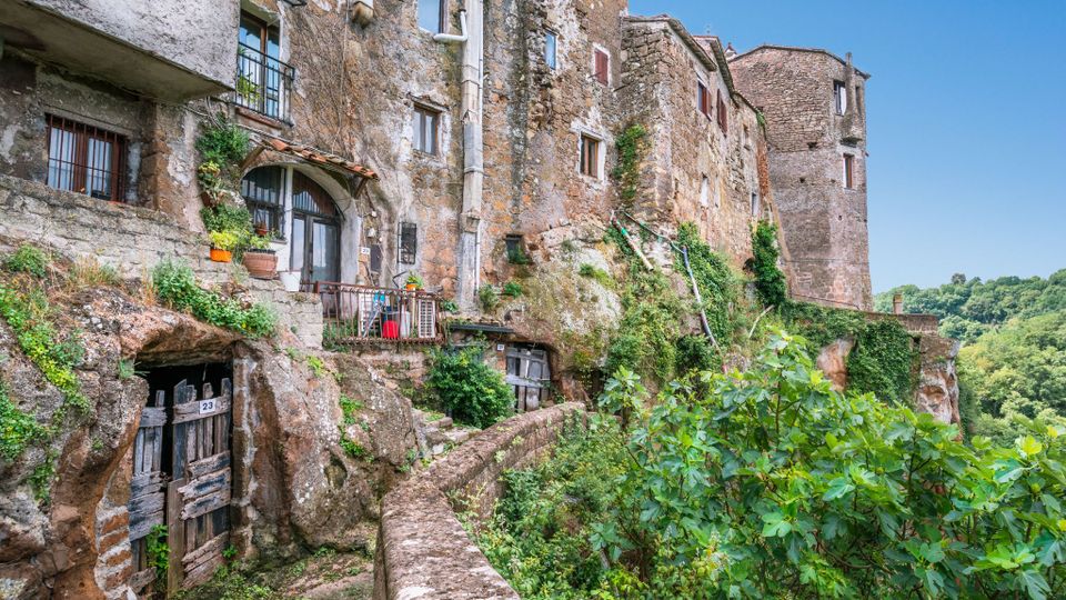 Borgo medievale con case in pietra addossate a una rupe, con facciate in muratura, finestre e archi, e porte ricavate nella roccia lungo un camminamento con muretto. Vegetazione rampicante e alberi circondano l’abitato, affacciato su una valle verde sotto un cielo sereno.