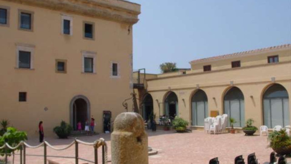 Cortile lastricato in mattoni tra edifici color crema, con un alto palazzo a più piani e un porticato ad archi con grandi vetrate sul lato destro. In primo piano si vede un pilastro in pietra e, lungo il perimetro, catene tra paletti, piante in vaso e alcune sedie impilate.