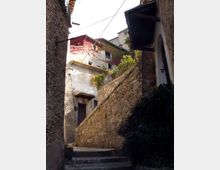 Vicolo stretto in un borgo storico, con scalini in pietra e un alto muro in muratura che sale verso case in intonaco e pietra. Si vedono persiane, un lampione a parete, tetti sporgenti e cavi sospesi tra gli edifici.