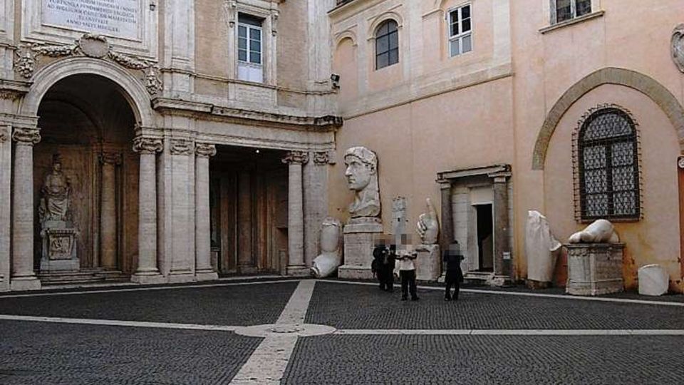 Cortile interno in pietra con pavimentazione a sampietrini, circondato da edifici storici in stucco chiaro. A sinistra si vede un portico con archi e colonne classiche con iscrizione monumentale, mentre lungo le pareti sono esposte grandi sculture e frammenti in marmo.