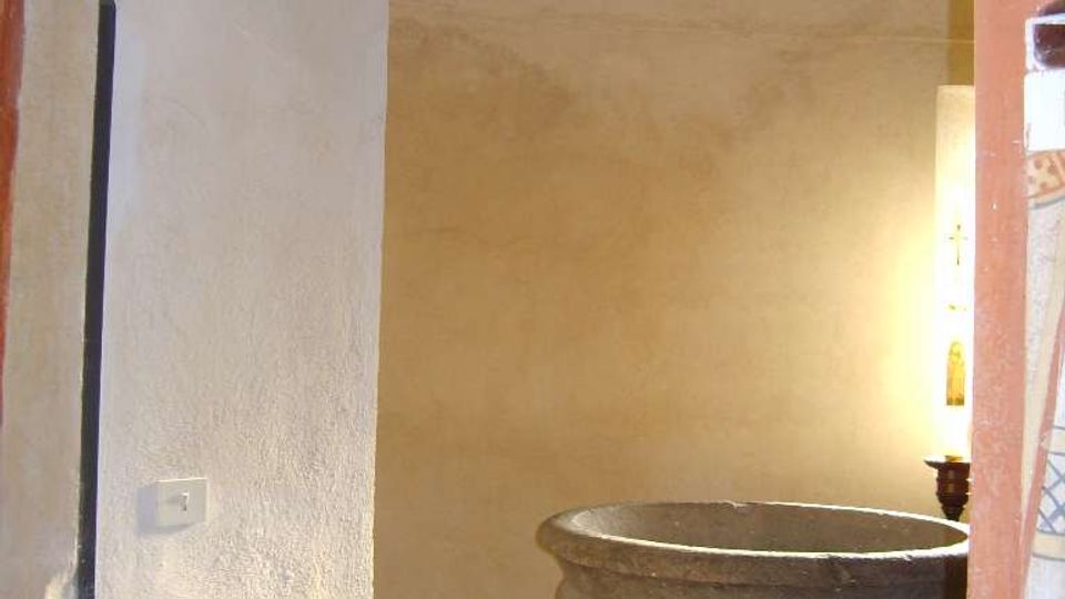 Interno di una piccola stanza con pareti intonacate chiare e pavimento in mattonelle, vista attraverso una porta. Al centro si trova una grande vasca circolare in pietra su base, illuminata da una lampada sul lato destro.