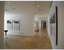 Interno di una galleria o museo moderno con pareti bianche e pavimento in grandi lastre di pietra, illuminato da faretti incassati nel soffitto. Lungo le pareti sono esposti pannelli informativi e fotografie, con un corridoio che si estende in profondità verso altre sale.