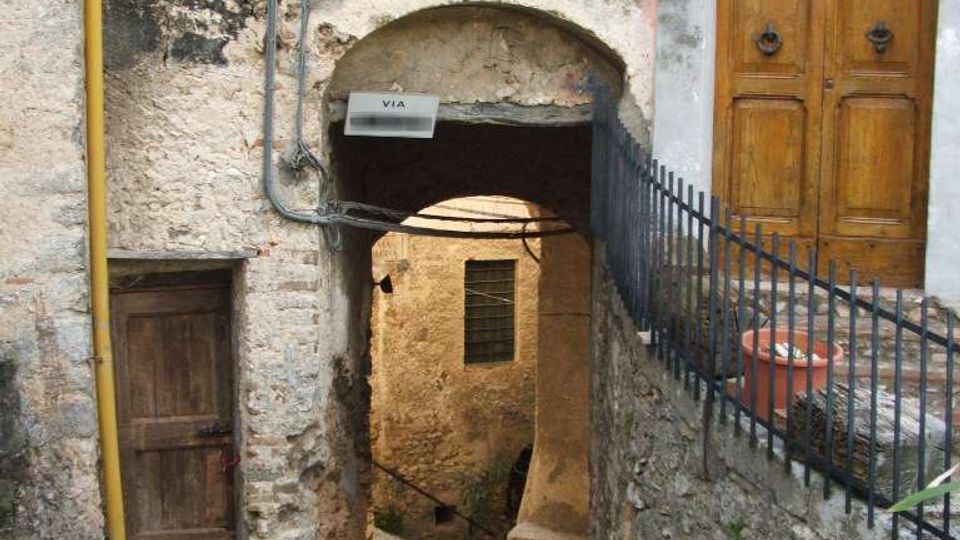 Stretto vicolo in pietra di un borgo antico, con passaggio ad arco sotto un edificio intonacato e scrostato e una piccola finestra con grata. Sulla destra si vede un portone in legno con arco superiore e una ringhiera in ferro lungo un muretto di pietra.