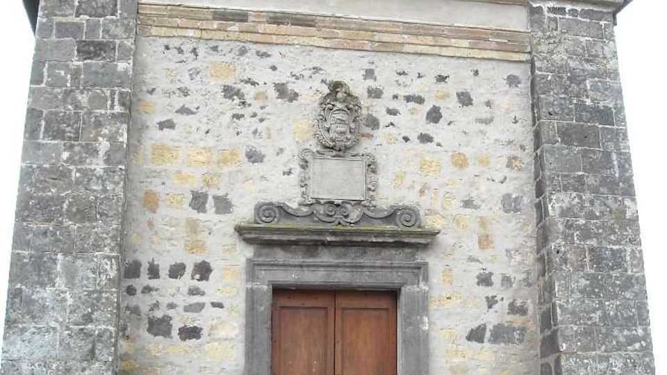 Facciata in pietra di una piccola chiesa o cappella storica con timpano triangolare e oculo circolare in alto. Al centro si vede un grande portale in legno incorniciato da pietra, con stemma decorativo sopra e due piccole finestre con inferriate ai lati.