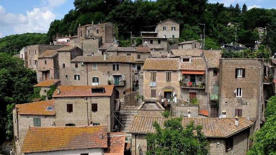 Borgo collinare con case in pietra addossate tra loro e tetti in coppi, disposte su più livelli con scalinate e piccoli balconi. Sullo sfondo una fitta area boschiva circonda l’abitato.