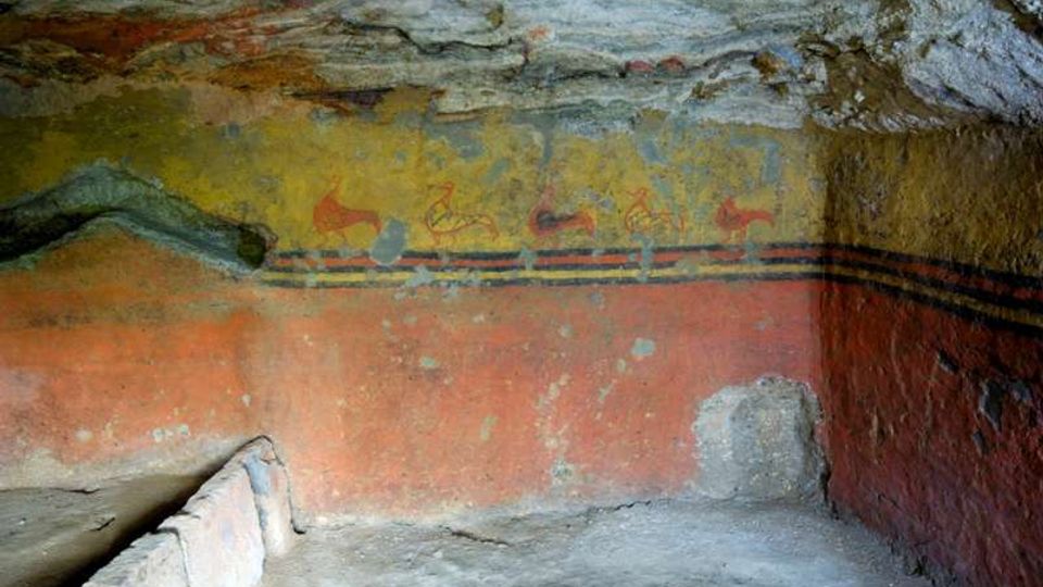 Interno di una tomba ipogea scavata nella roccia, con pareti dipinte da affreschi in giallo e rosso con fasce decorative e figure stilizzate. Sul lato sinistro si vede una banchina in pietra e il soffitto roccioso grezzo sopra la camera.