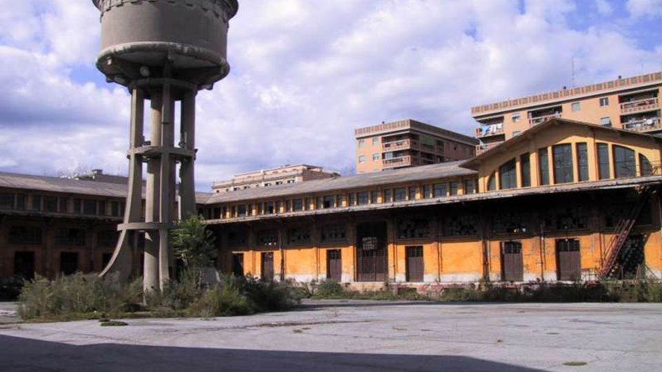 Grande cortile industriale con un alto serbatoio d’acqua cilindrico su pilastri in cemento e un edificio basso a pianta semicircolare con facciata arancione e finestre ad arco. Sullo sfondo si vedono palazzi residenziali e cielo parzialmente nuvoloso.