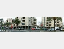 Ampia strada urbana con incrocio e strisce pedonali, fiancheggiata da palme e palazzi residenziali multipiano con balconi. Lungo i marciapiedi sono presenti auto parcheggiate e segnaletica stradale, tra cui cartelli di divieto di accesso.
