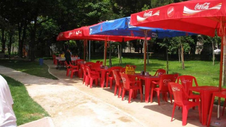 Area ristoro all’aperto in un parco, con tavoli e sedie di plastica rossi disposti lungo un vialetto curvo. Una fila di grandi ombrelloni rossi e blu con loghi Coca‑Cola copre i posti a sedere, circondati da prato e alberi.